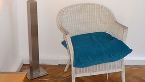 Een witte rotan stoel met een blauwgroen kussen, een hoge vloerlamp met verlichting, en een schilderij van blauwe bloemen aan de muur.