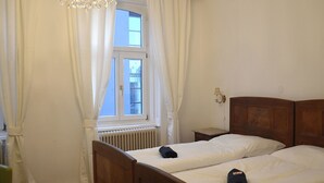 Een slaapkamer met twee houten bedden, witte gordijnen, een radiator, een klein nachtkastje en een kroonluchter.