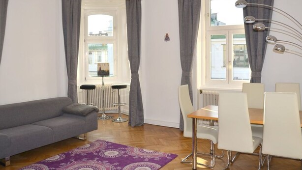 Een moderne woonkamer met een grijze bank, een eettafel met stoelen, een kroonluchter en uitzicht op de stad door grote ramen met grijze gordijnen.