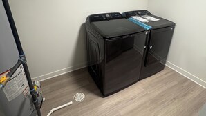 Et vaskerum med en sort vaskemaskine og tørretumbler, trægulv og en ventilationsrist på væggen.