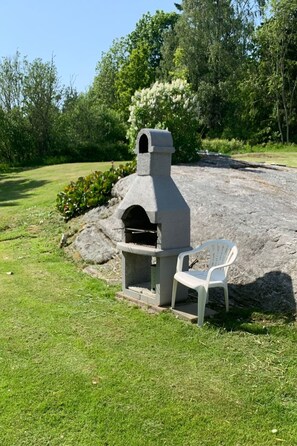 Una parrilla de piedra al aire libre con chimenea, una silla de plástico blanco y un césped con árboles de fondo.