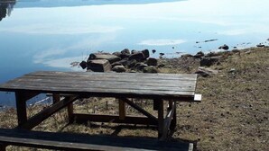 A wooden picnic table by a calm lake with rocks and sparse vegetation.