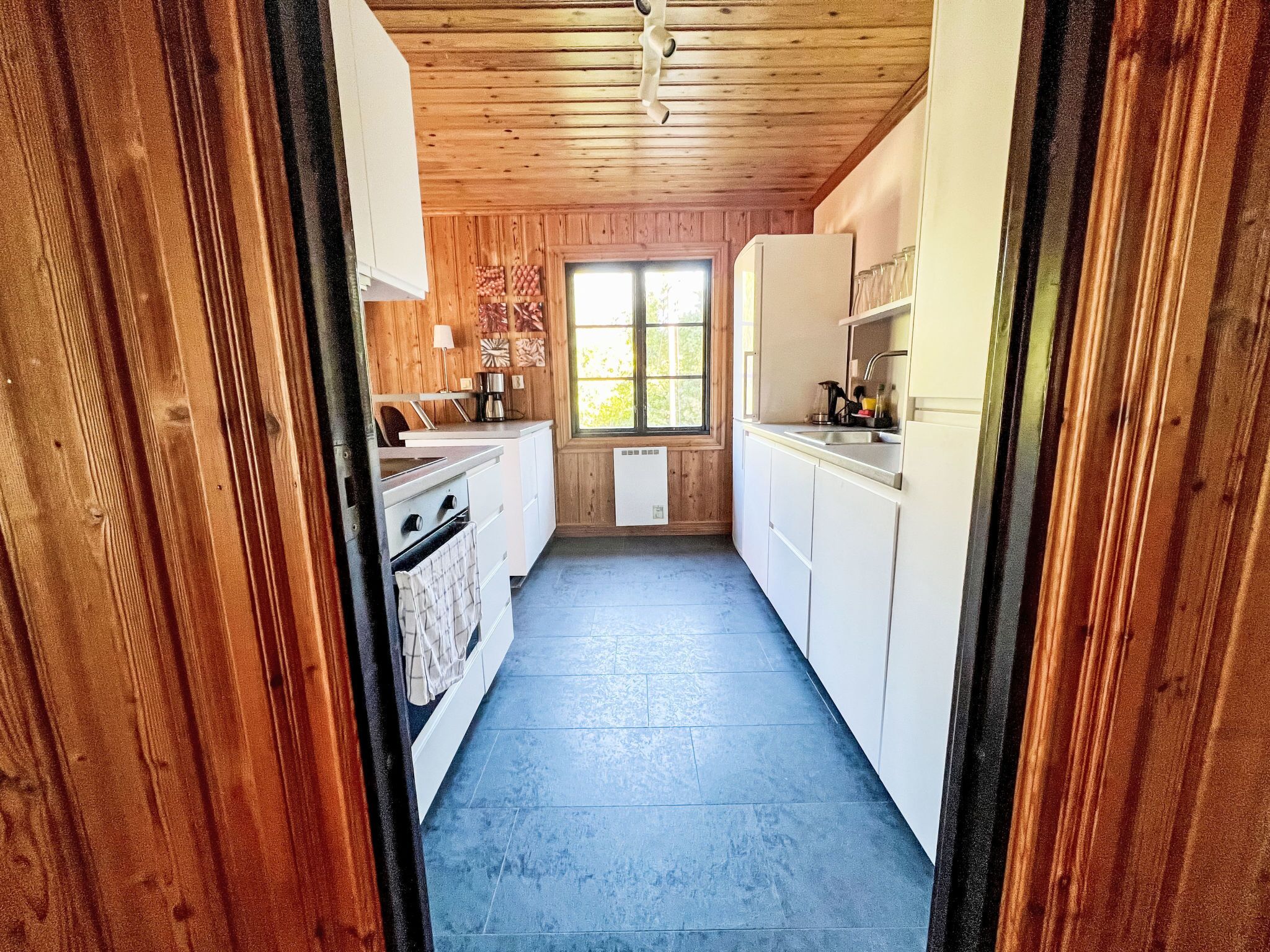 A narrow kitchen with wooden walls, a window, and a blue floor.