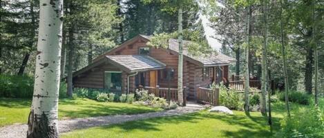 A log cabin with a porch, surrounded by trees and a grassy yard.