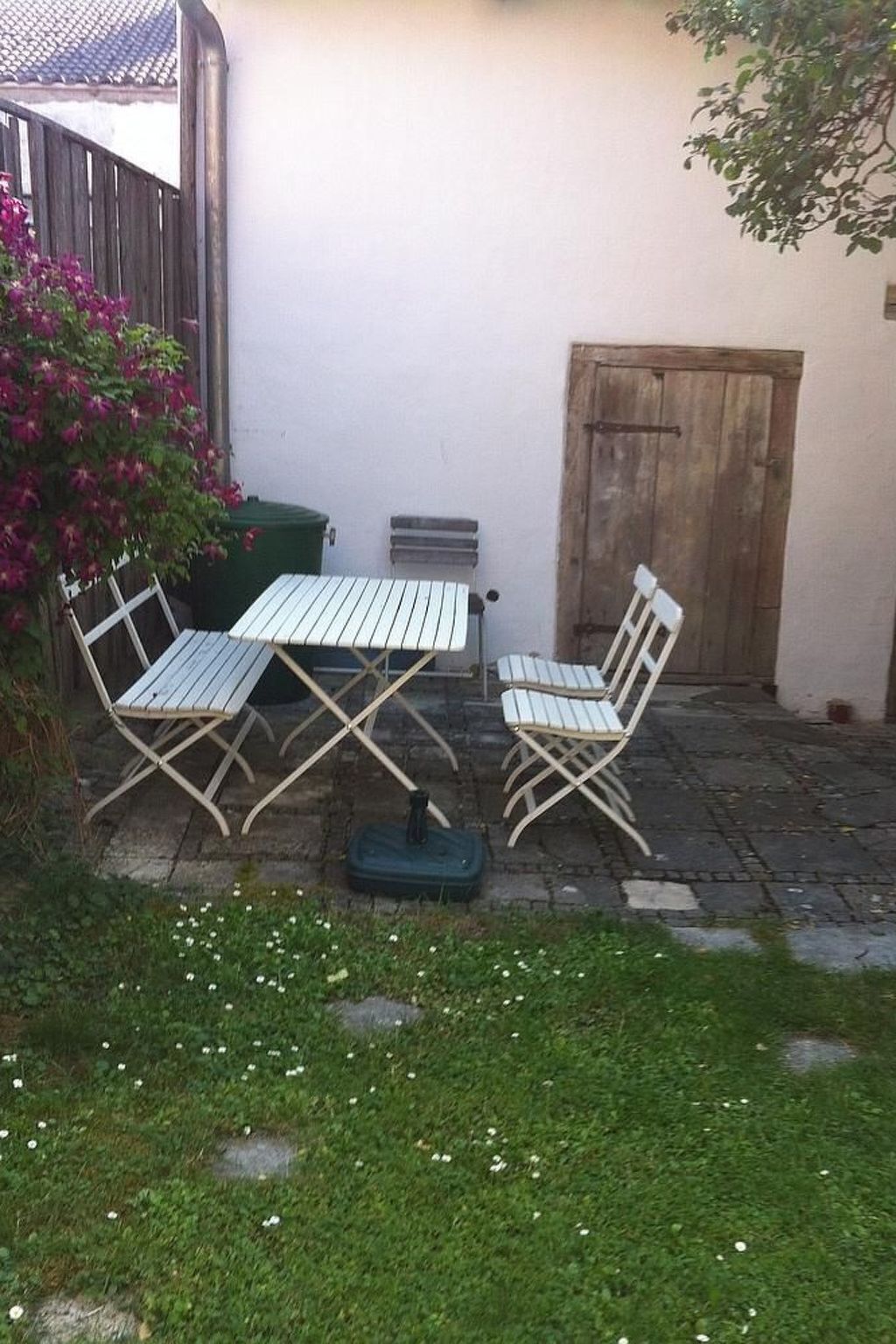 Een klein buitenterras met een tafel en twee stoelen, een groene prullenbak en een houten deur.
