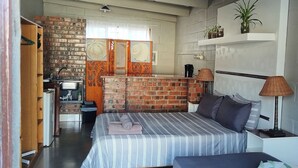 Quarto com parede de tijolos, cama com roupa de cama listrada, uma pequena escrivaninha, abajur e prateleira com plantas.