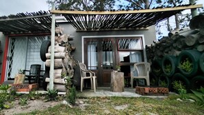 Uma casa com varanda coberta, uma pilha de lenha e diversos vasos com plantas e móveis de jardim.