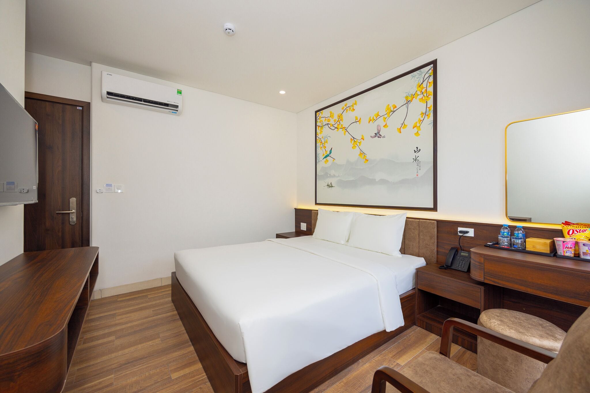 A hotel room with a large bed, a wooden desk, a chair, a bedside table, a mirror, and a wall-mounted air conditioner.