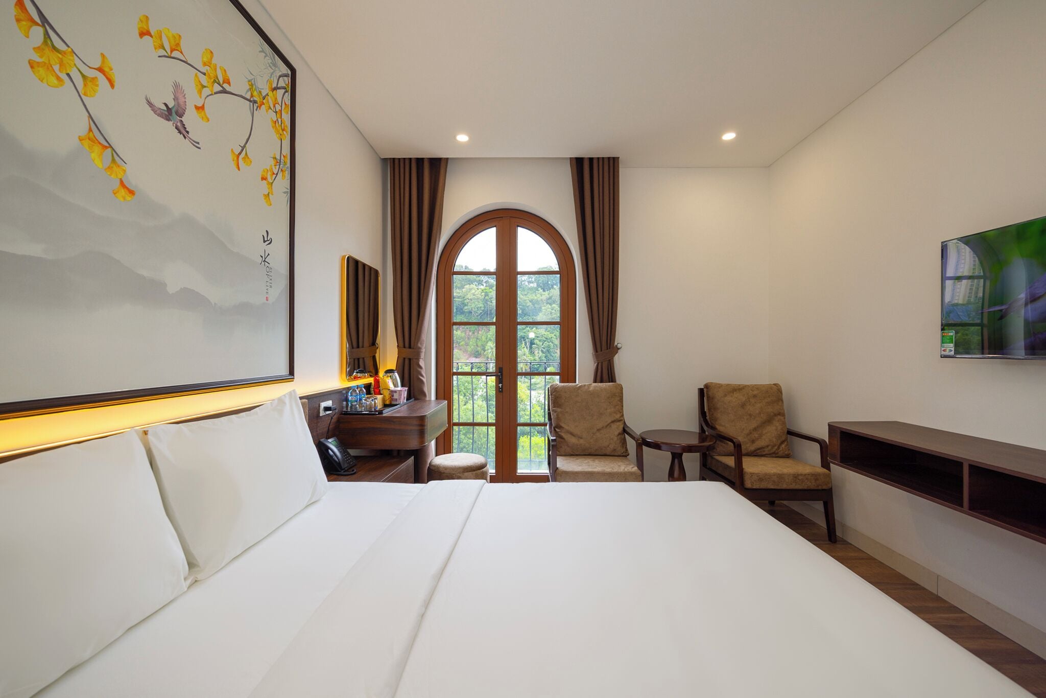 A hotel room with a large bed, a desk with a lamp, a chair, a TV mounted on the wall, and a large window with curtains.