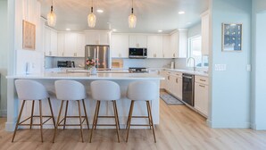 A modern kitchen with white cabinets, a central island with bar stools, stainless steel appliances, and pendant lighting. - Pacific Edge  Private Beach Access, Oceanfront Luxury Retreat 4BR 3BA (Smith River)