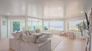 A spacious living room with a large gray sectional sofa, a wooden coffee table, and a view of the ocean through large windows. - Pacific Edge  Private Beach Access, Oceanfront Luxury Retreat 4BR 3BA (Smith River)