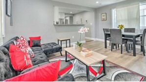 Una sala de estar moderna con un sofá gris, cojines rojos y una mesa de centro de madera. Hay un comedor con una mesa y sillas, y una cocina al fondo.