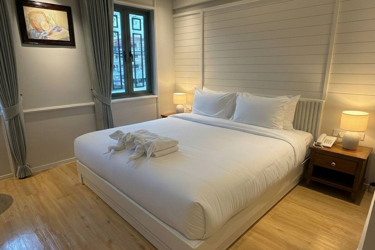 Un lit bien fait, avec du linge de lit et des serviettes blanches, une lampe de chevet et un tableau encadré au mur.