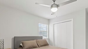 Un dormitorio con cama, mesita de noche, ventilador de techo, ventana con persianas y una puerta blanca.