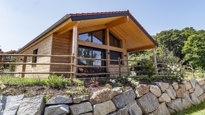 A wooden cabin with a sloped roof, large windows, and a stone wall in front. - Komfortables Ebenerdiges Chalet im Vital Camp Bayerbach (Bayerbach)