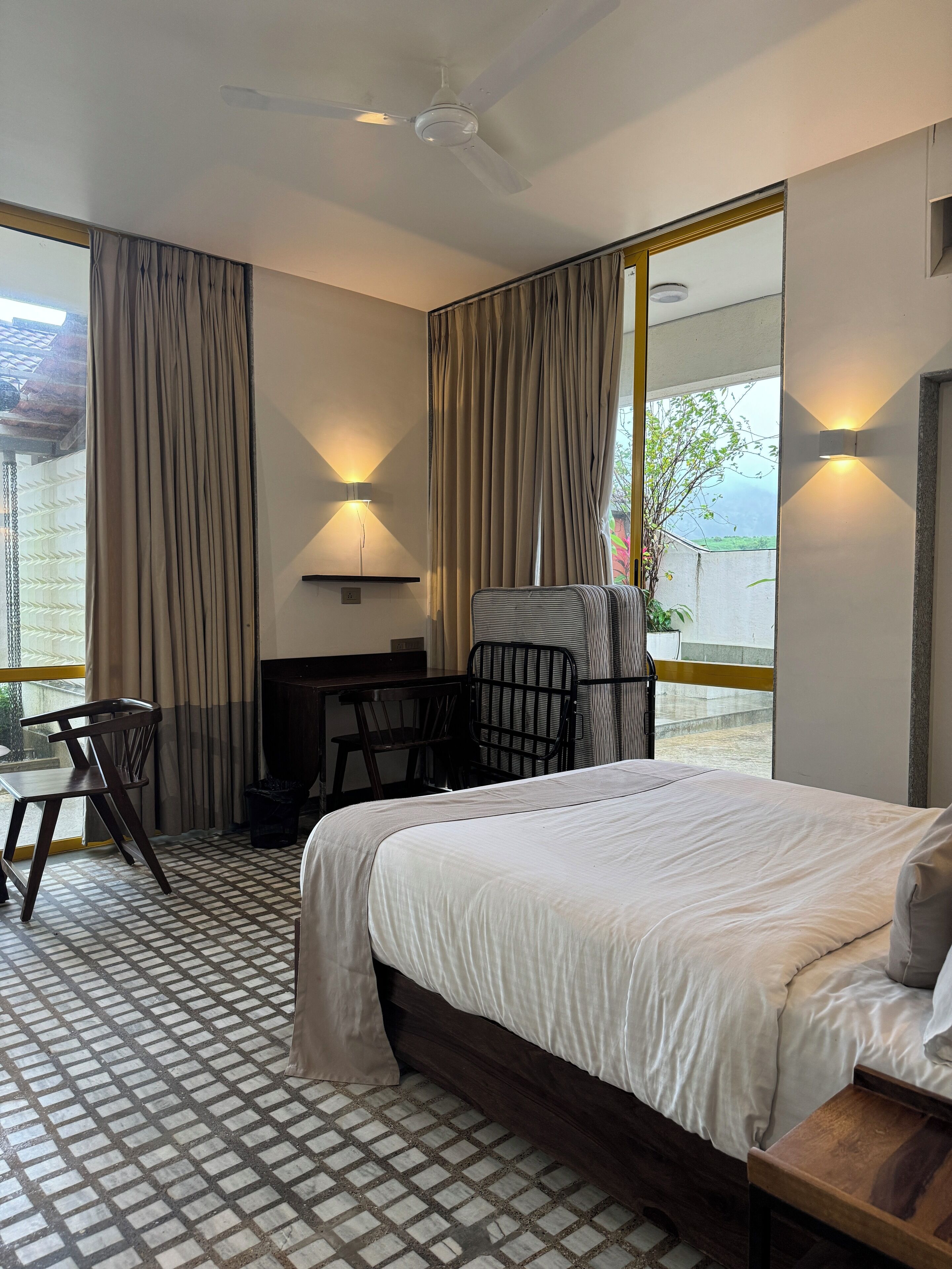 A hotel room with a bed, a desk, a chair, and a small table. There are curtains on the windows and a ceiling fan.