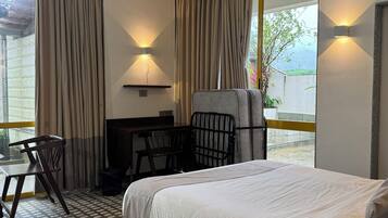 A hotel room with a bed, a desk, a chair, and a small table. There are curtains on the windows and a ceiling fan.