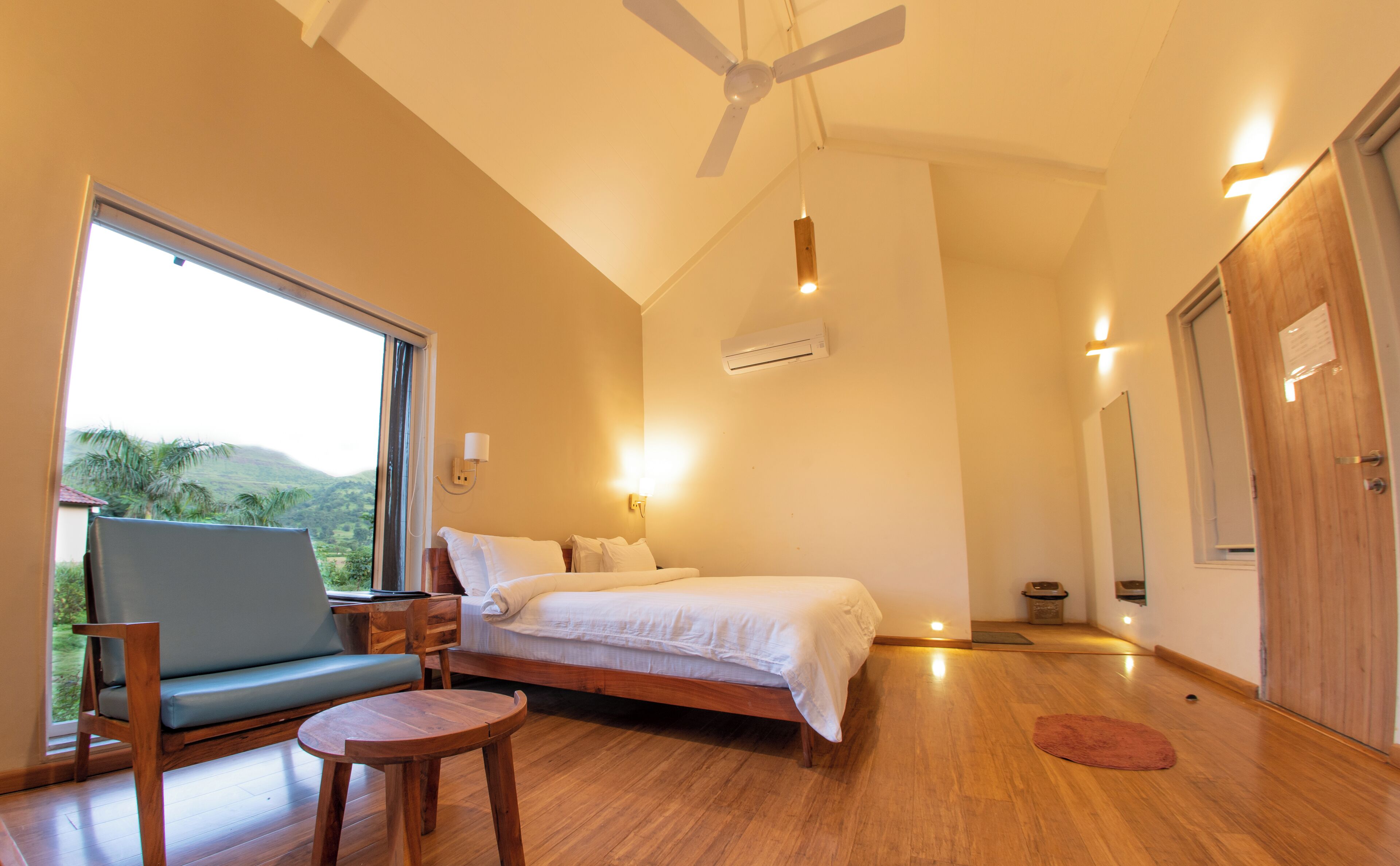 A bedroom with a bed, a chair, a small table, a ceiling fan, and a window with a view of greenery.