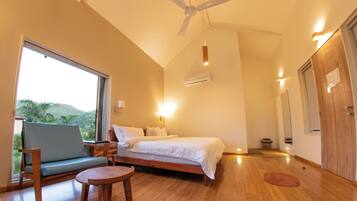 A bedroom with a bed, a chair, a small table, a ceiling fan, and a window with a view of greenery.