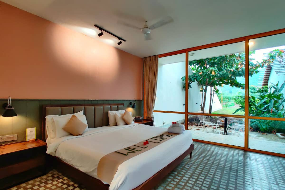 A hotel room with a large bed, a nightstand, a lamp, and a view of the outdoors through a large window.