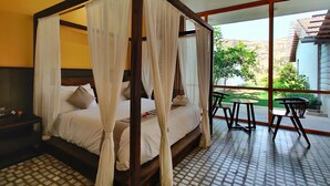 A four-poster bed with white linens and a canopy, a small table with chairs, and a view of a garden and building through large glass doors. - Touchwood Bliss Resort - Igatpuri (Igatpuri)