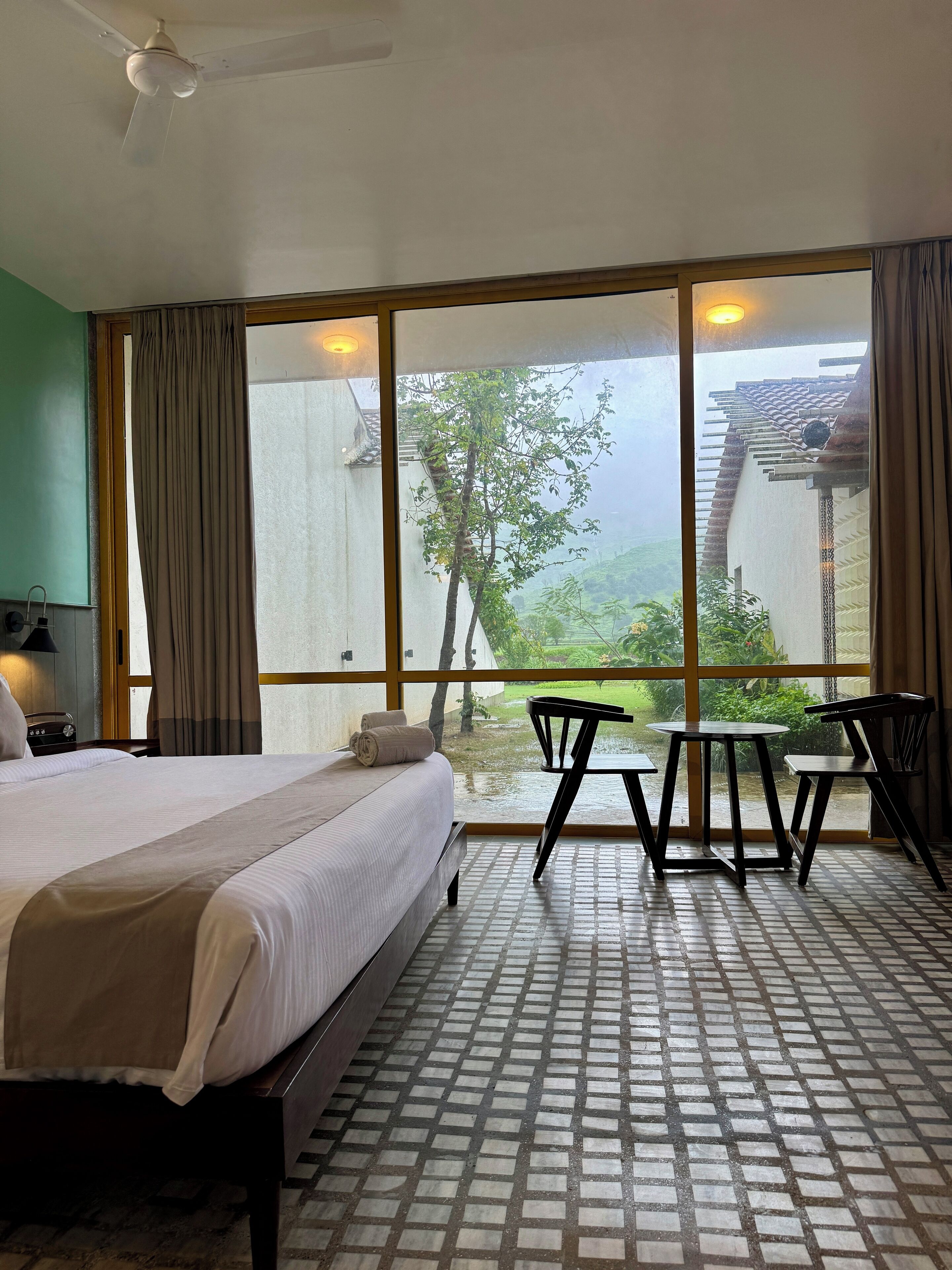 A hotel room with a bed, a table, and chairs, large windows with a view of greenery, and a ceiling fan.