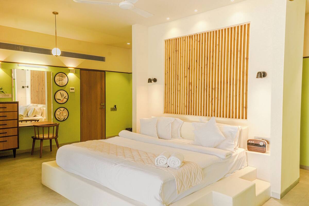 A modern bedroom with a large bed, a wooden slat headboard, a desk with a chair, and a mirror.