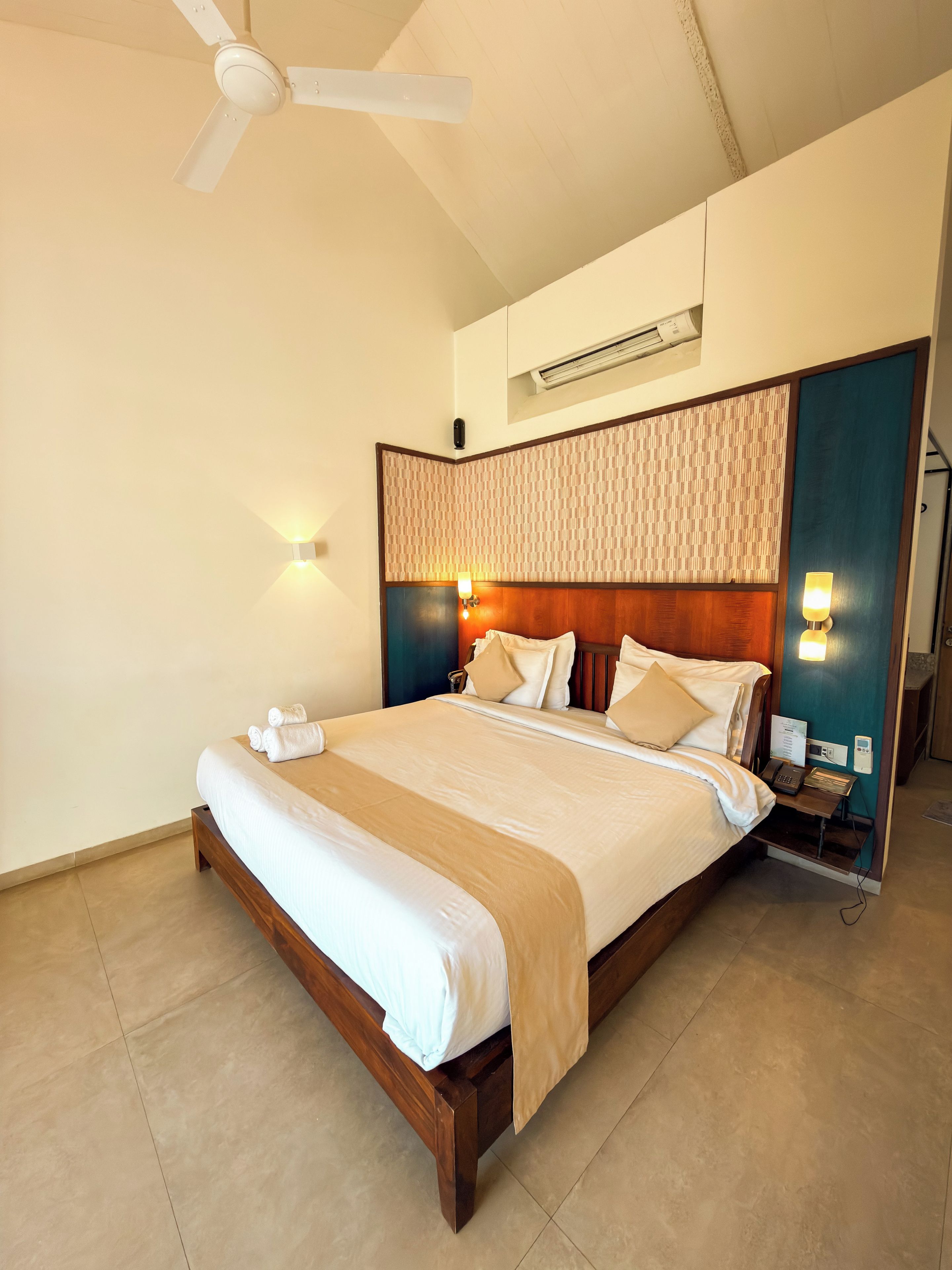 Habitación de hotel con una cama grande, ventilador de techo, aire acondicionado de pared y mesita de noche con teléfono y lámpara.