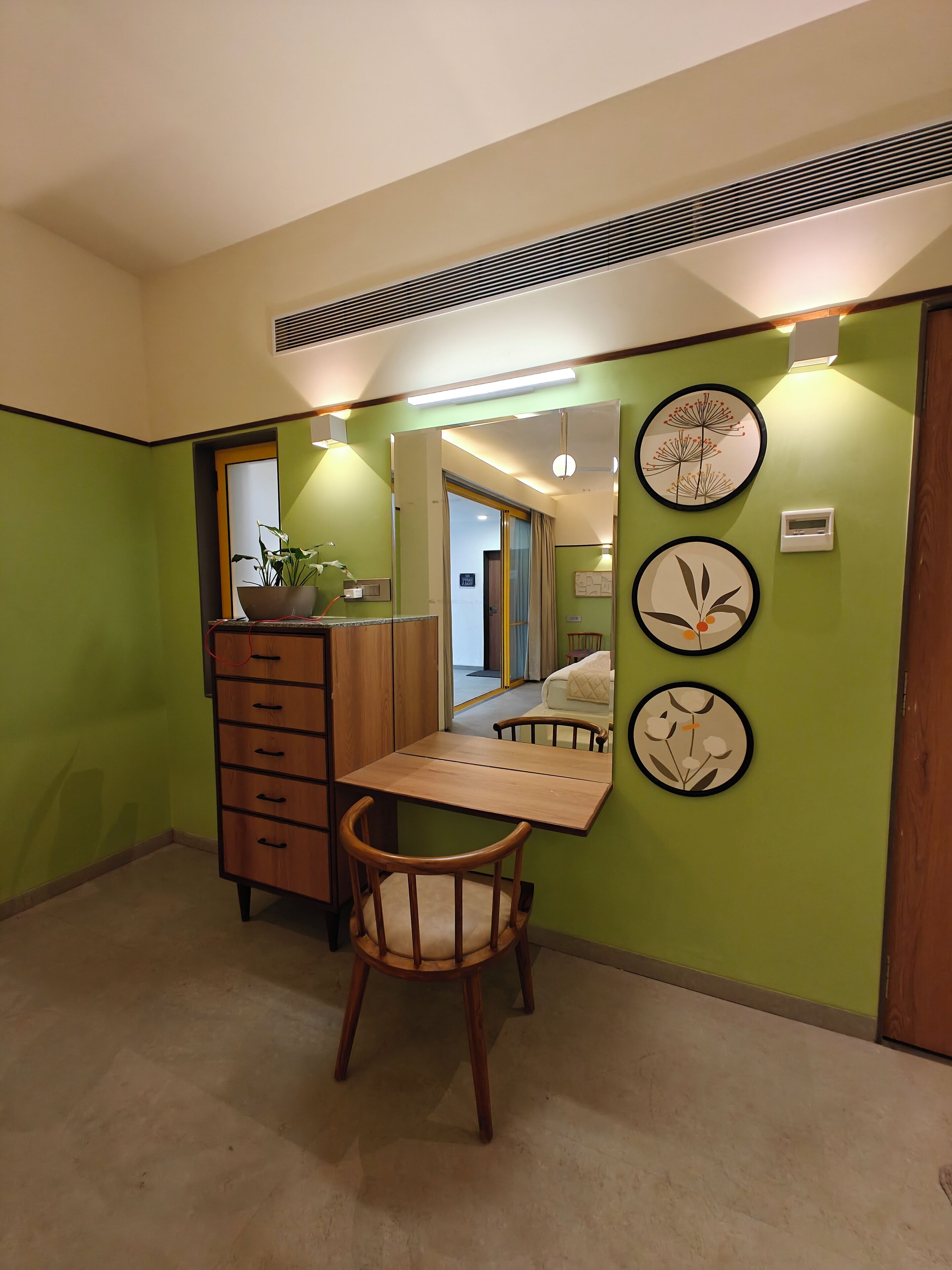 Quarto com cômoda de madeira, uma cadeira, escrivaninha e espelho. Há decorações nas paredes e uma ventilação no teto.