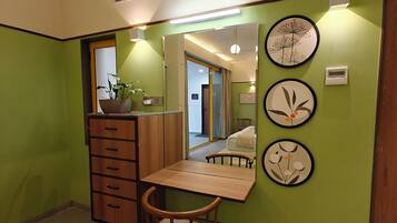 Quarto com cômoda de madeira, uma cadeira, escrivaninha e espelho. Há decorações nas paredes e uma ventilação no teto.