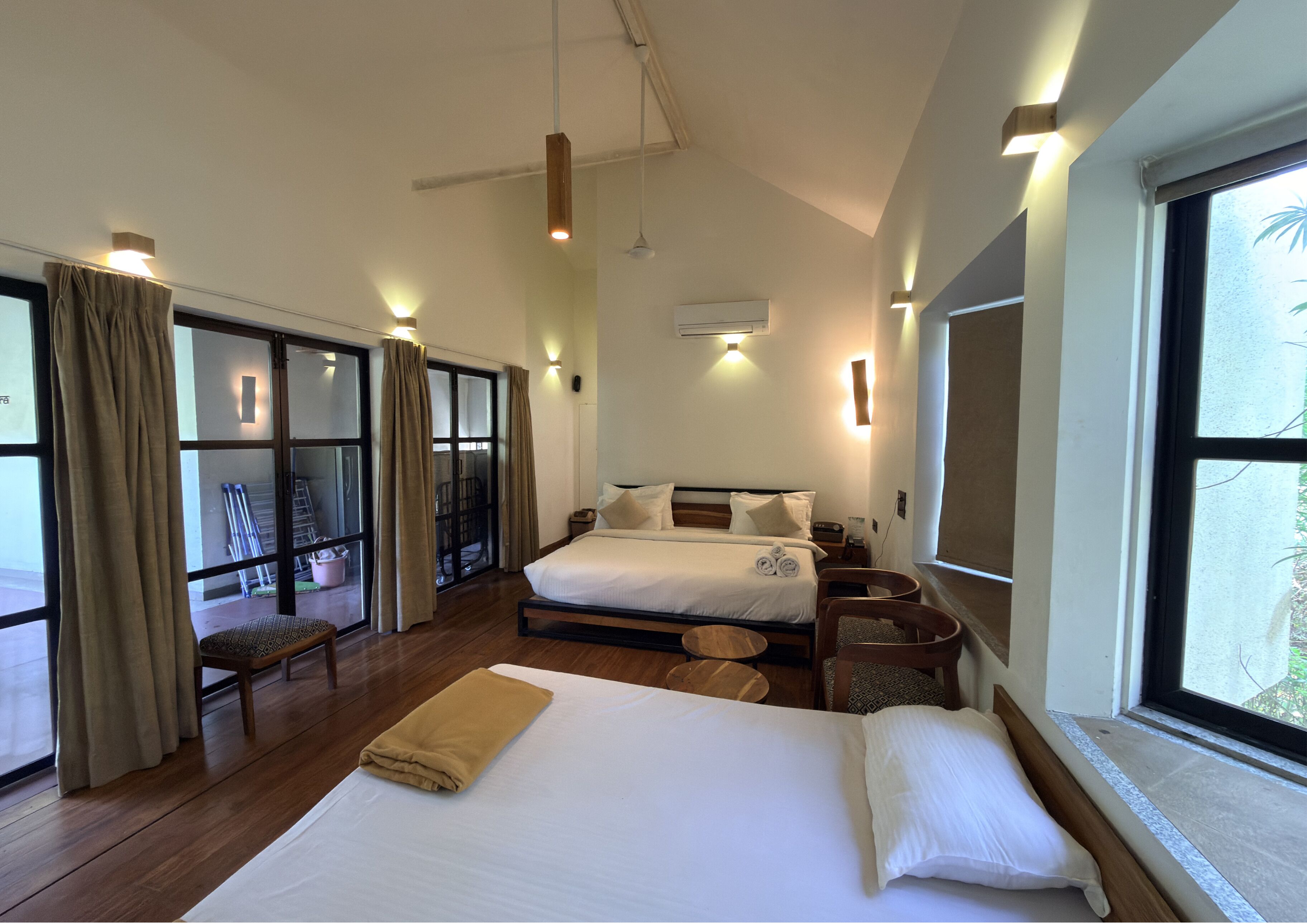 A hotel room with a large bed, a balcony with curtains, a wooden floor, and a view of greenery outside.