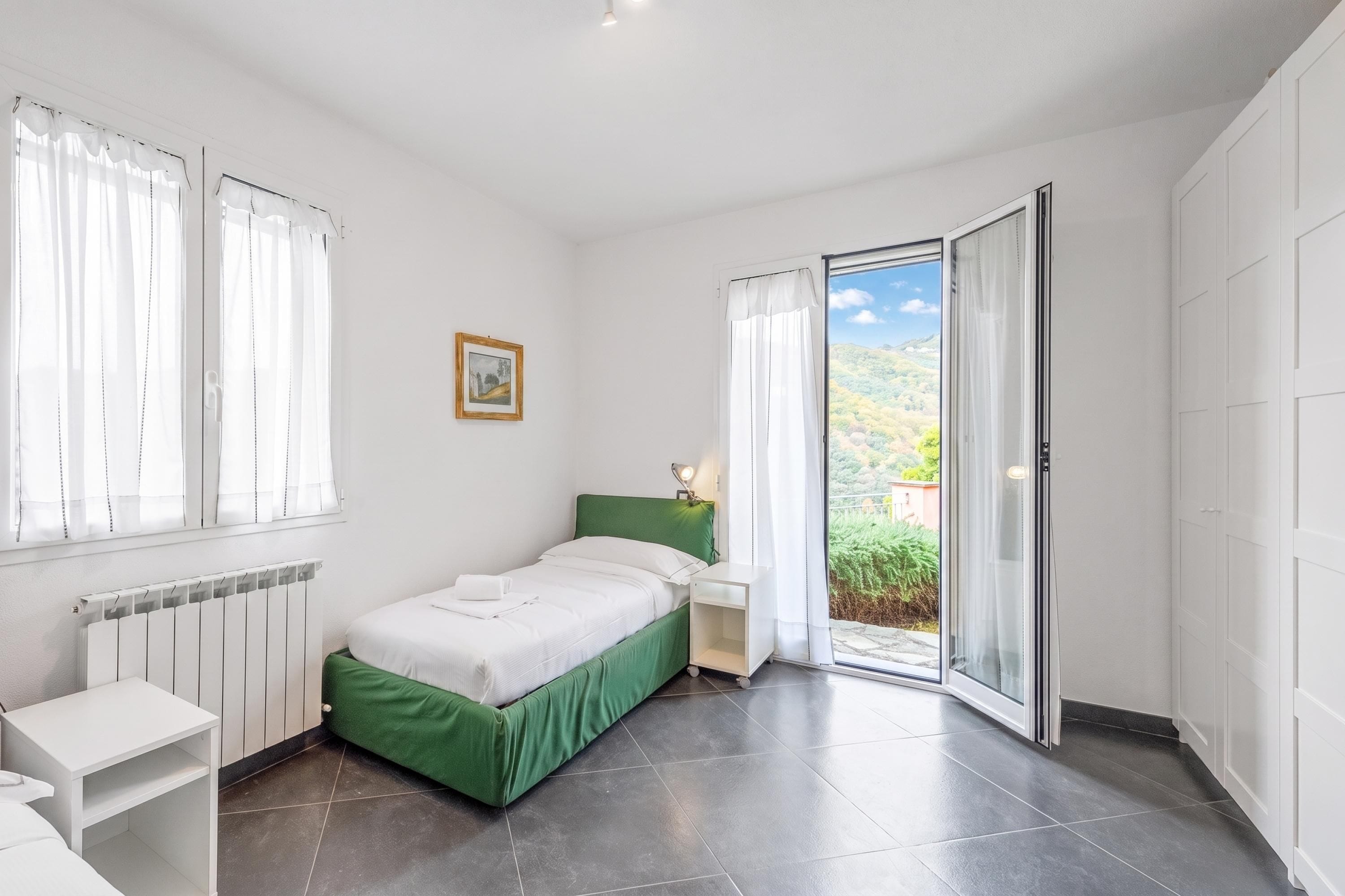 Une chambre à coucher comprenant un lit, une table de chevet, un radiateur, une fenêtre avec des rideaux et une vue sur la verdure à l’extérieur.