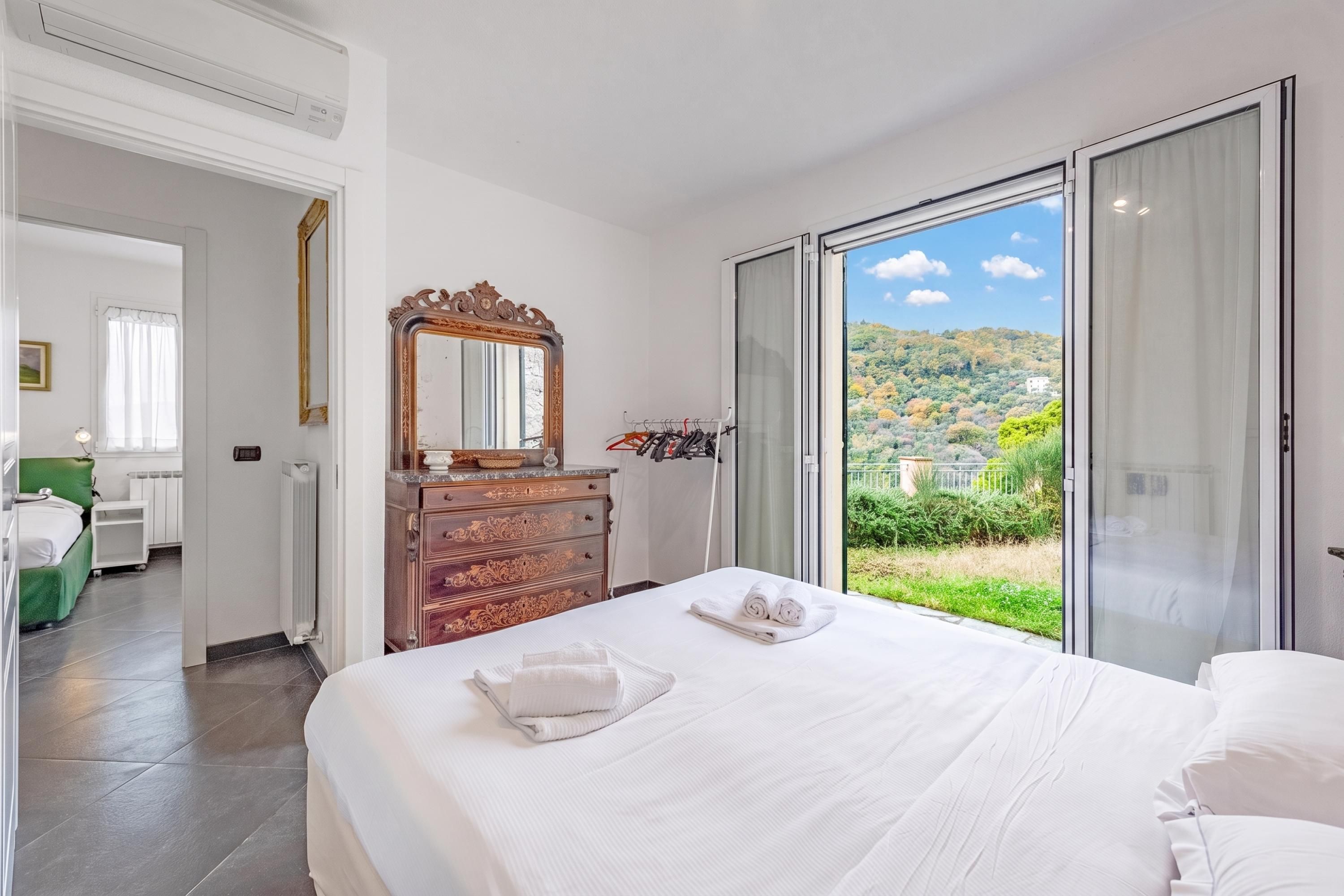 A hotel room with a large bed, a dresser, a mirror, and a view of a green landscape through a sliding glass door.