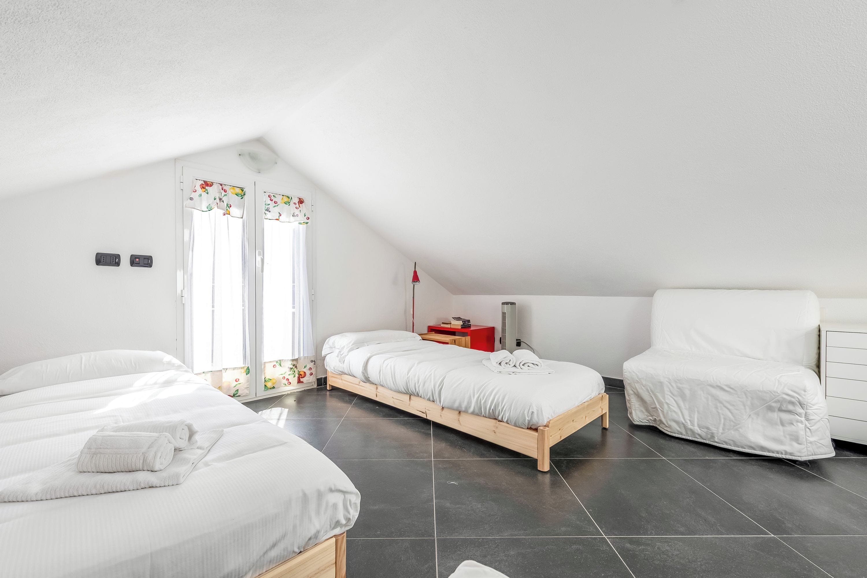 Un dormitorio con techo inclinado, dos camas, una mesita de noche, una silla y una ventana con cortinas.