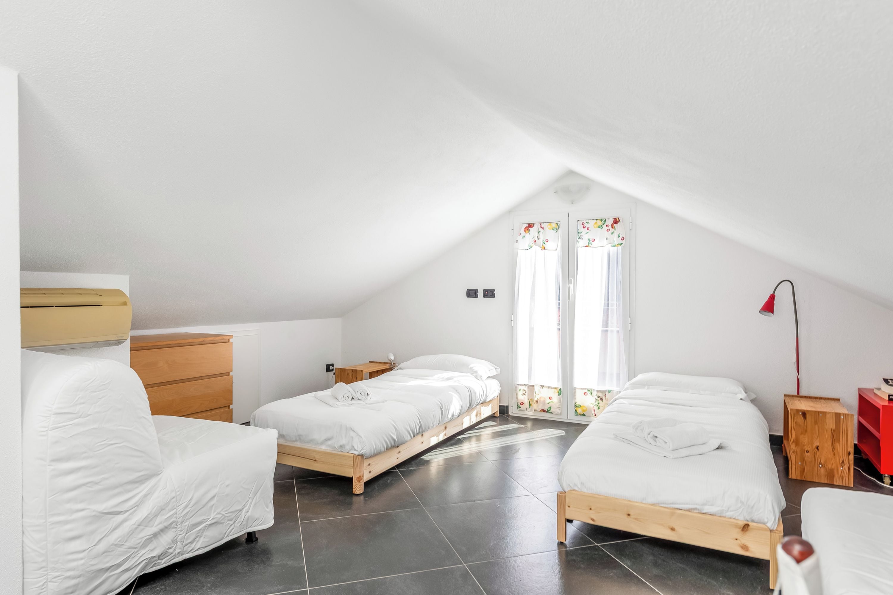 Una habitación con techo inclinado, dos camas individuales, un aparador de madera, una lámpara roja y una ventana con cortinas.