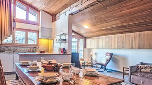 Un comedor rústico con una mesa de madera dispuesta para una comida, una cocina al fondo y una zona de estar acogedora con un sillón de cuero.