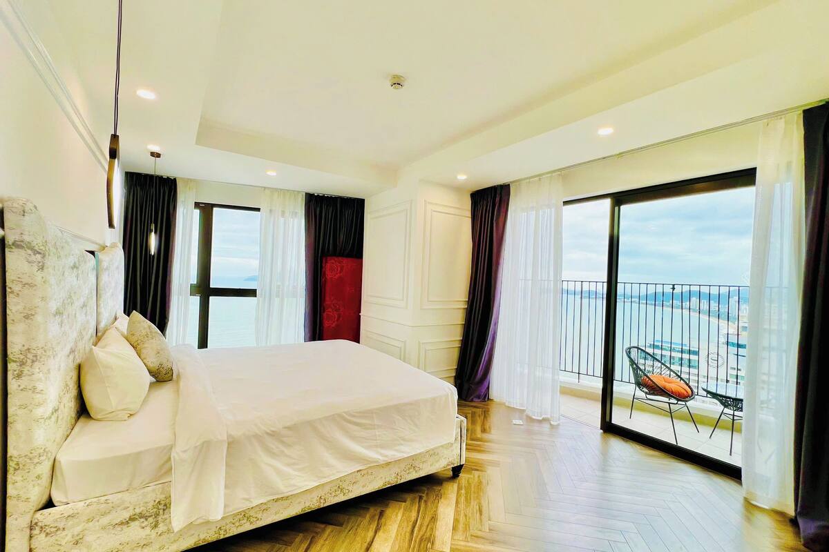 A spacious bedroom with a large bed, a balcony with a view of the sea, and a wooden floor.