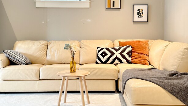 Un salon moderne avec un canapé en cuir beige, une petite table d’appoint en bois et un vase contenant des fleurs séchées. Deux œuvres d’art abstraites encadrées ornent le mur.
