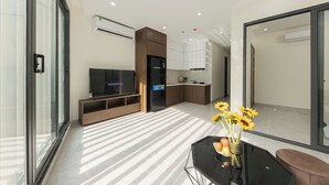 Un salon moderne avec un téléviseur à écran plat, une table basse en verre ornée d’un vase de fleurs, et un coin cuisine en arrière-plan.