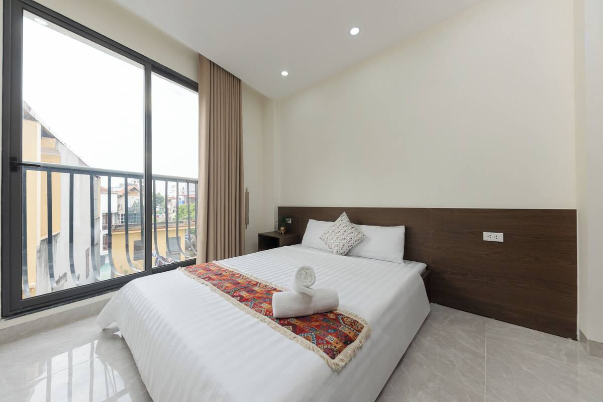 A hotel room with a large bed, a nightstand, a window with curtains, and a view of buildings outside.