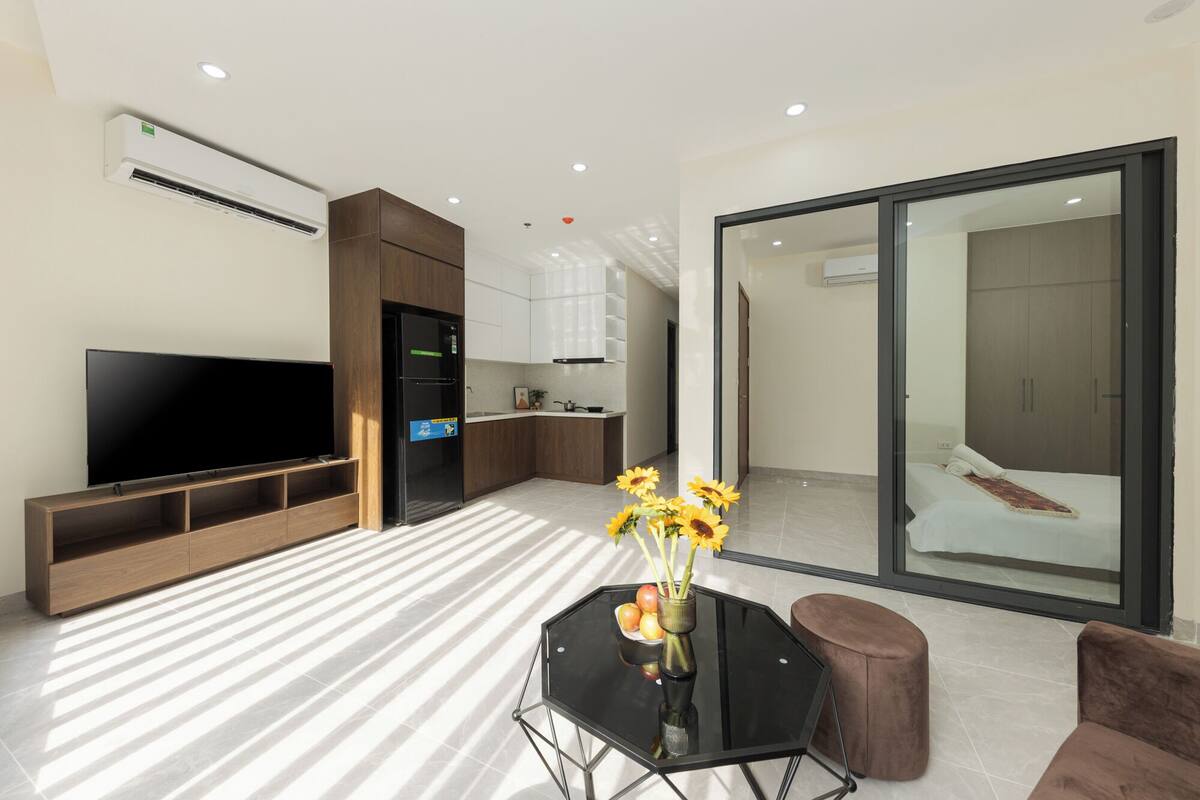 A modern living room with a flat-screen TV, a dining table with a vase of flowers, and a sliding glass door leading to a bedroom.