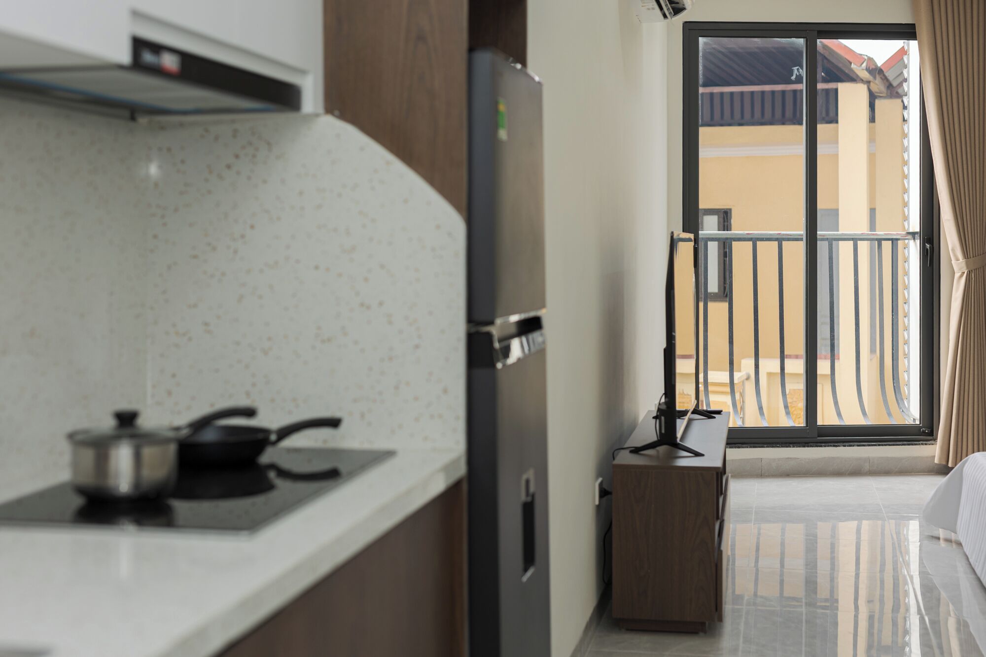 A hotel room with a kitchenette, a refrigerator, a small wooden cabinet, and a balcony with a metal railing.