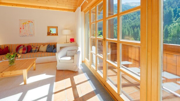 A spacious living room with a wooden ceiling, a large window with a forest view, a sofa with colorful cushions, a white armchair, and a wooden coffee table. - Talschluss 500 by Interhome (Saalbach-Hinterglemm)