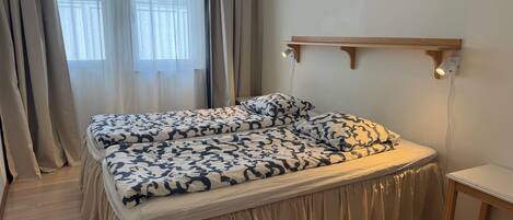 A double bed with a patterned bedspread, a bedside table, a wall-mounted light, and a window with sheer curtains.