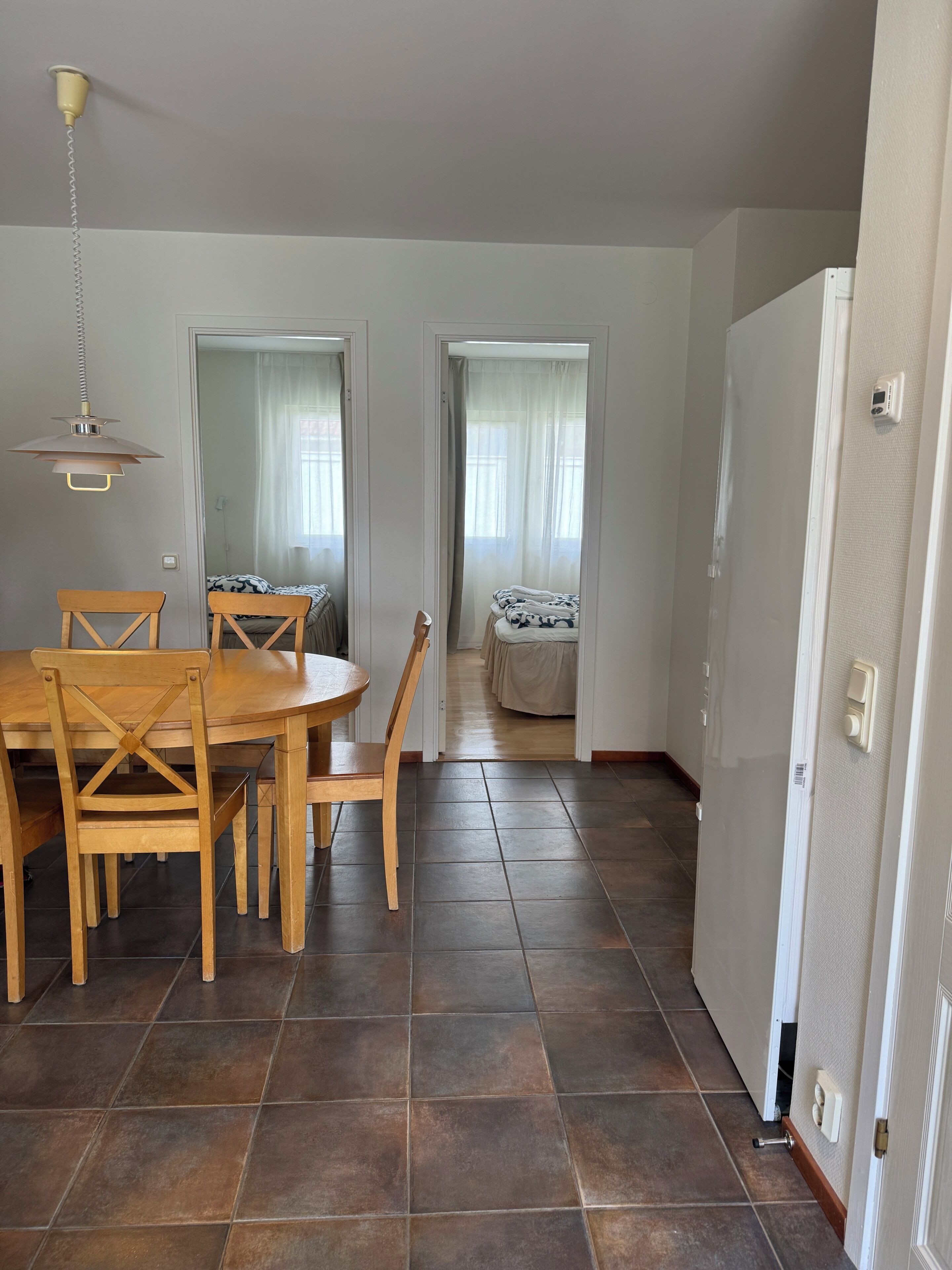 Un comedor con una mesa y sillas de madera, una lámpara colgante y vistas hacia un dormitorio con cama y cortinas.