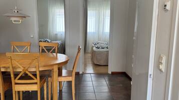 Un comedor con una mesa y sillas de madera, una lámpara colgante y vistas hacia un dormitorio con cama y cortinas.