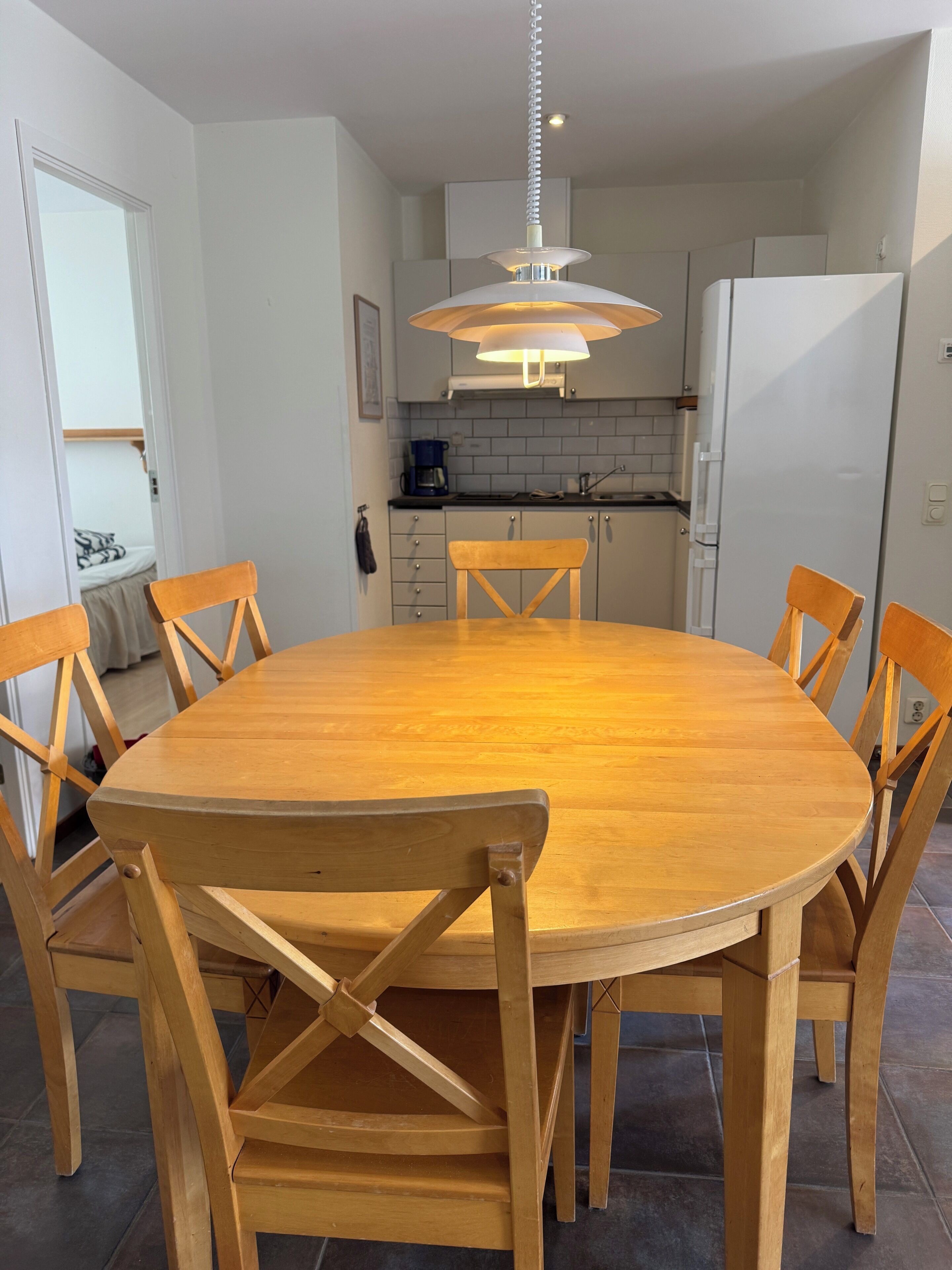 A round wooden dining table with six chairs, a hanging pendant light, and a kitchen in the background.