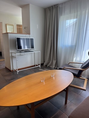 A modern room with a wooden oval table, two chairs, a television on a white cabinet, and a large window with sheer curtains.