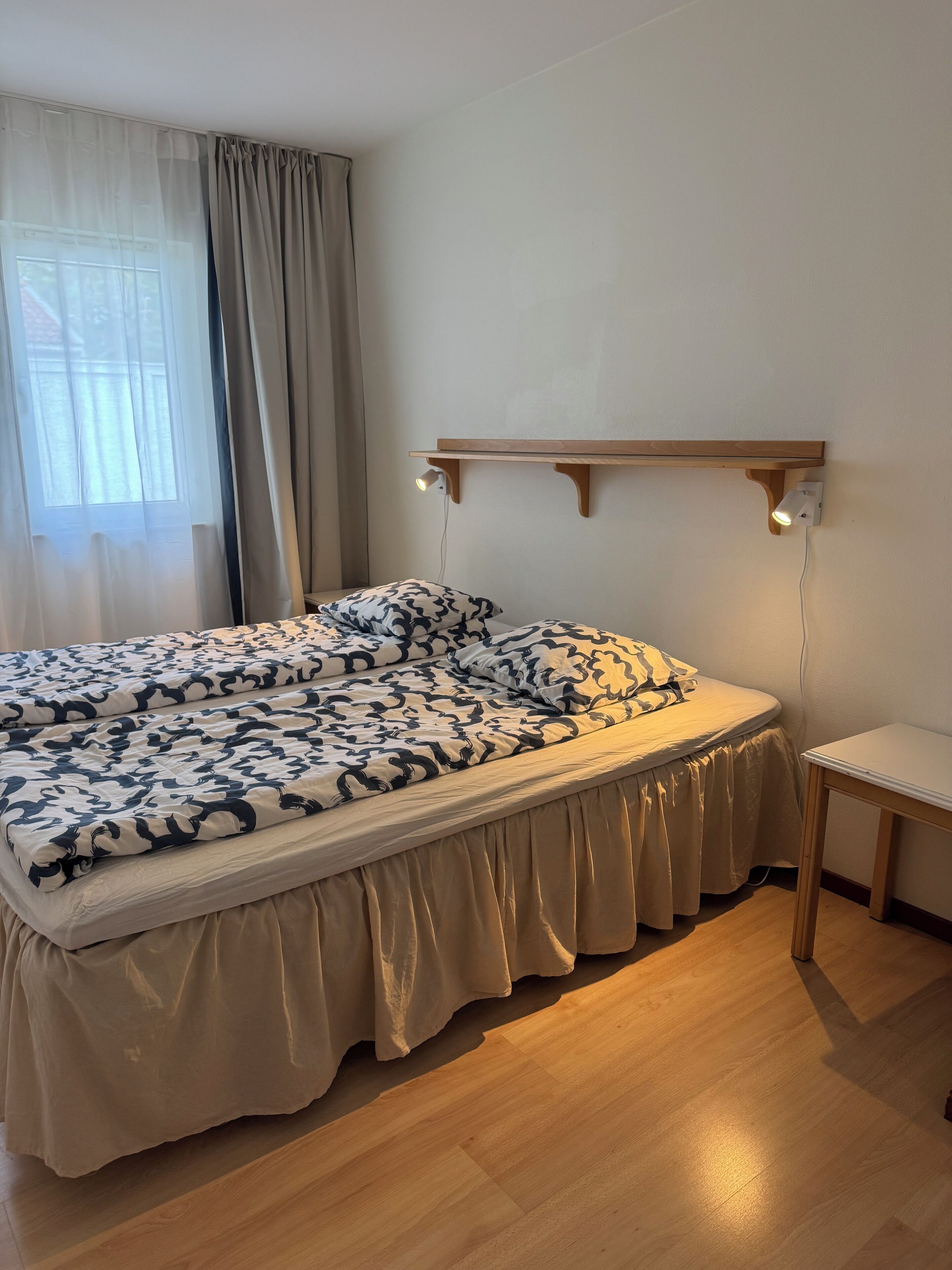 Une chambre à coucher avec un lit, une table de chevet, une étagère fixée au mur et une fenêtre avec des rideaux.