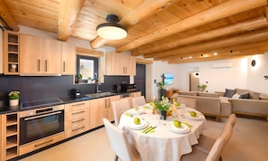 A modern kitchen with wooden cabinets, a dining area with a round table set for four, and a living area with a sofa and TV.
